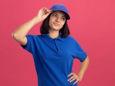 dissatisfied young delivery girl in blue uniform and cap looking aside puzzled standing over pink background
